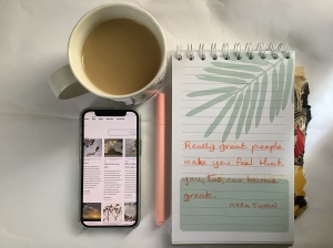 Smartphone showing blog posts, pastel pen, Mark Twain’s written on notebook, and coffee with milk on white background.