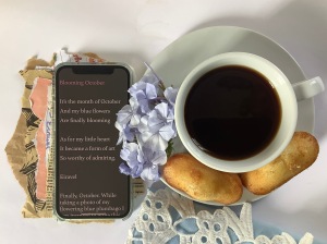 poem on smartphone beside black coffee in white cup, blue flowers, and two small madeleines cake.